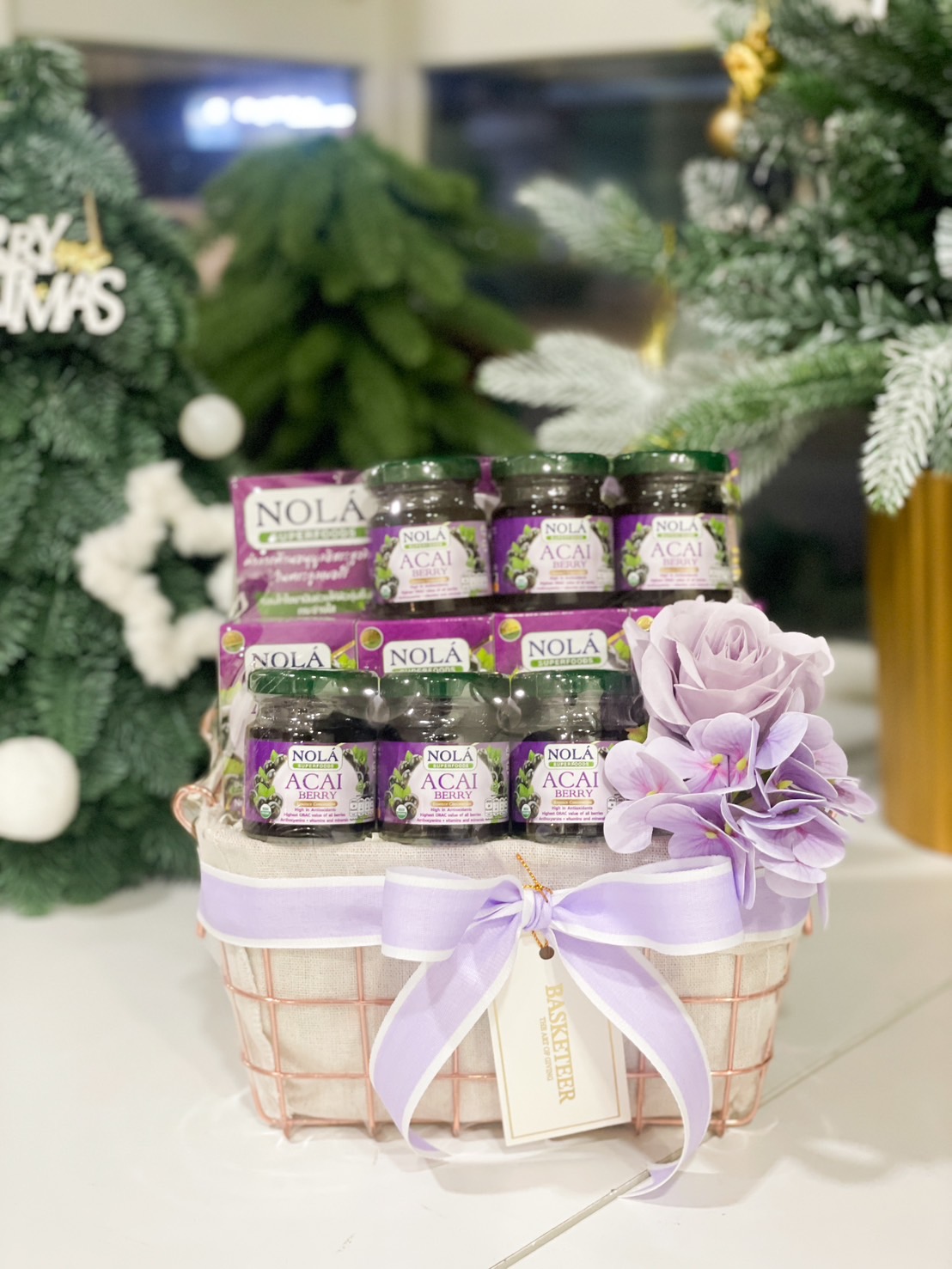 A pink wire basket filled with Nola Acai Berry drinks, adorned with a lilac bow and floral decorations, perfect for gifting.