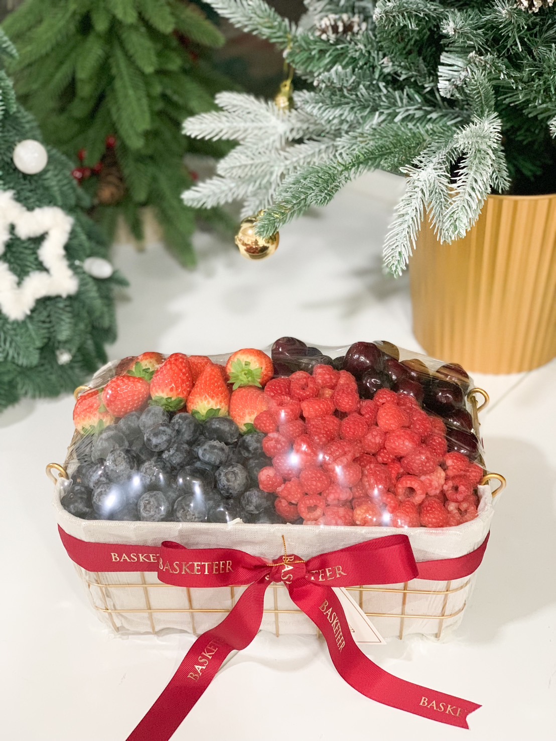 A golden fruit basket filled with fresh strawberries, blueberries, raspberries, and cherries, elegantly tied with a red ribbon.