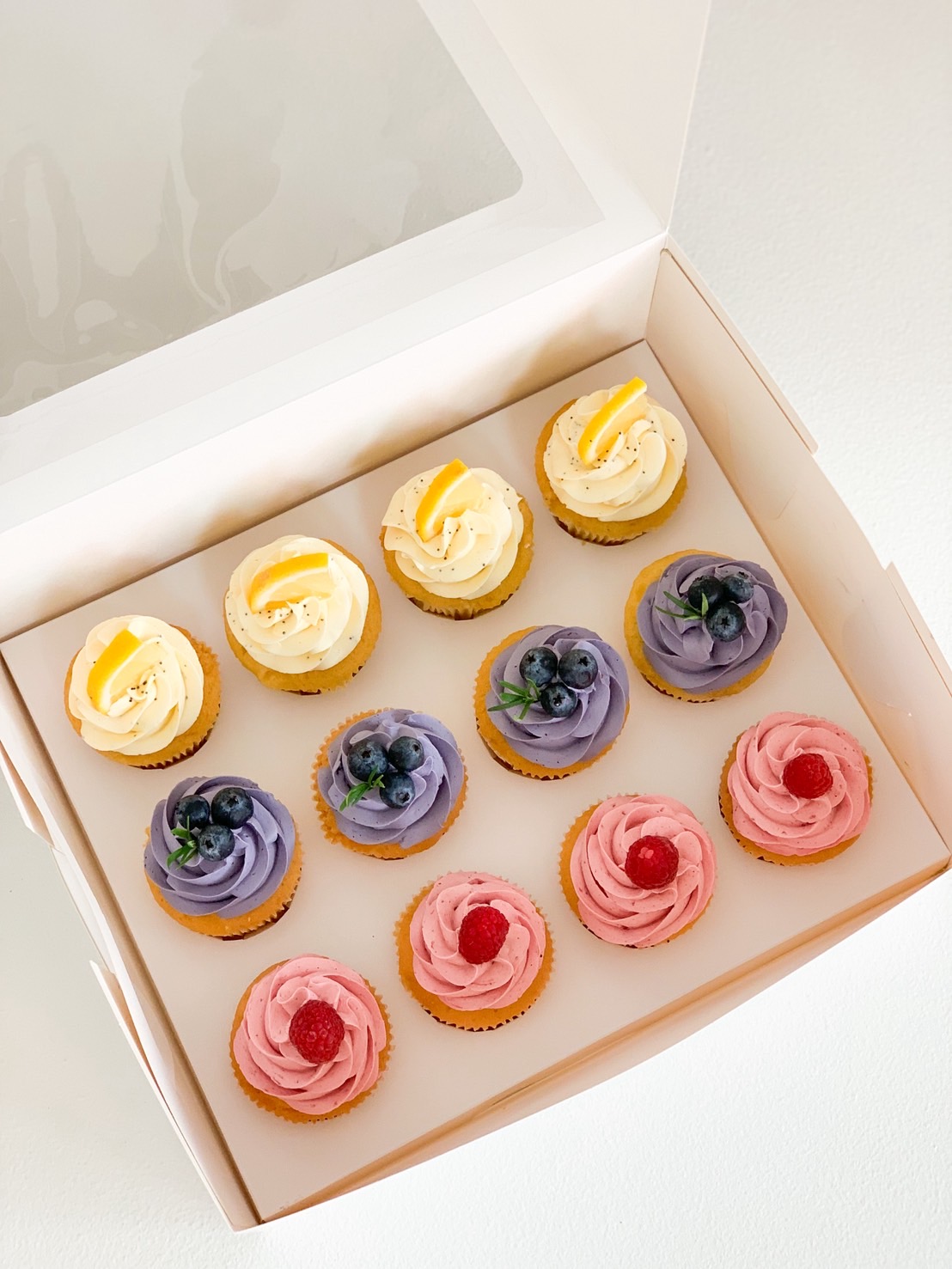 Box of assorted premium cupcakes with lemon, blueberry, and raspberry buttercream frosting in white gift box.