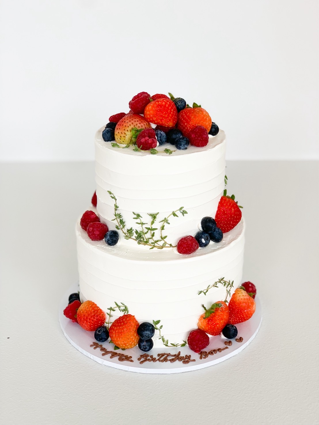 Two-tier white buttercream cake decorated with fresh strawberries, raspberries, blueberries, and thyme.