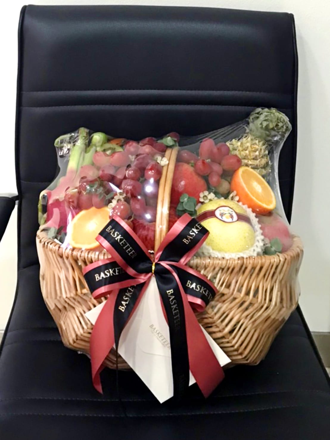 A wicker basket filled with a variety of orchard fresh fruits, including grapes, strawberries, an orange, and a pineapple, wrapped in clear cellophane. The basket is adorned with a black and red ribbon labeled 