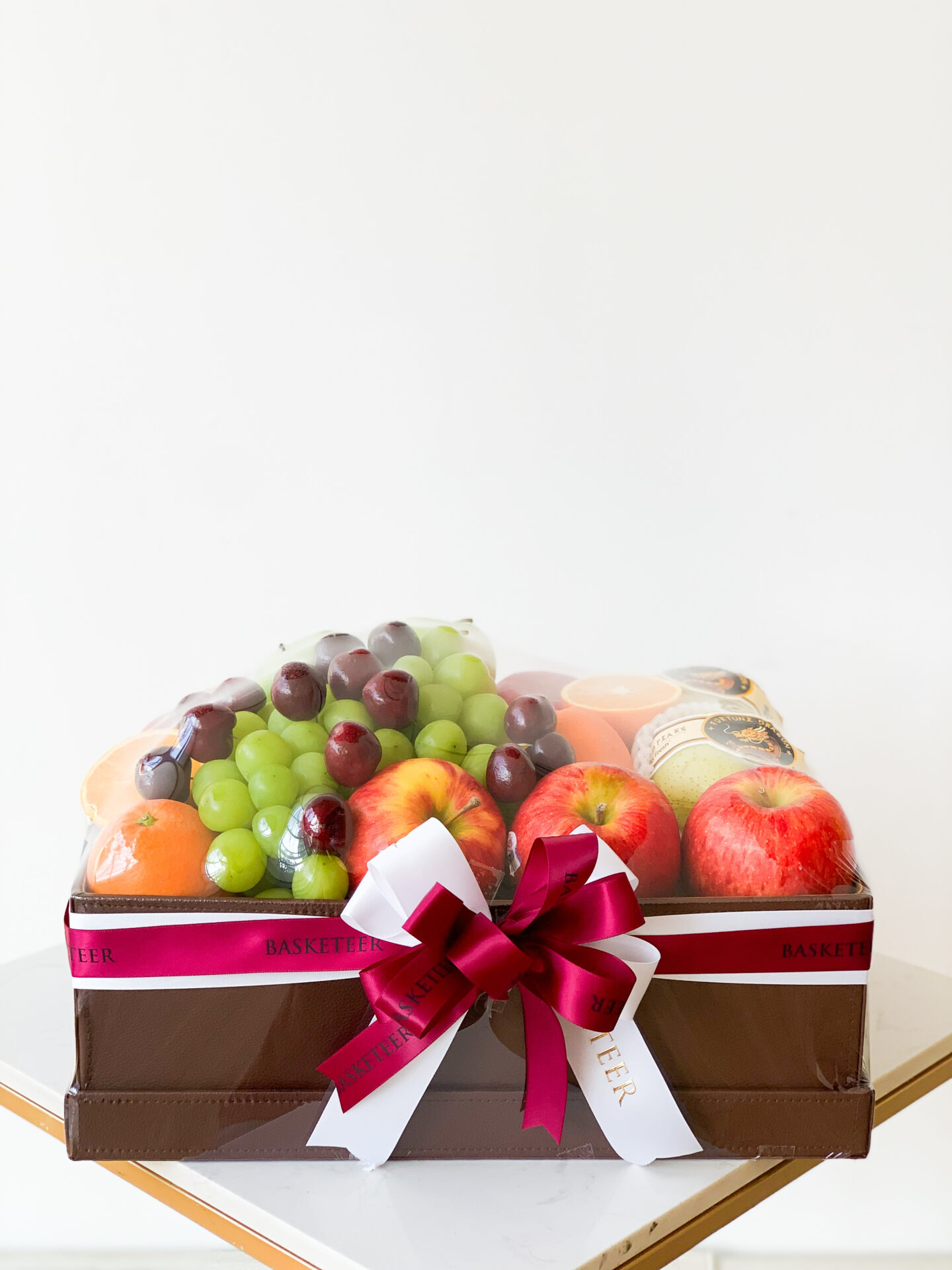A neatly wrapped gift basket brimming with green grapes, red grapes, red apples, oranges, and various jars. Tied with a white and dark pink ribbon displaying the BASKETEER brand name on both the ribbon and the brown box. This Premium Fruit Box Hamper sits elegantly on a white and gold table.