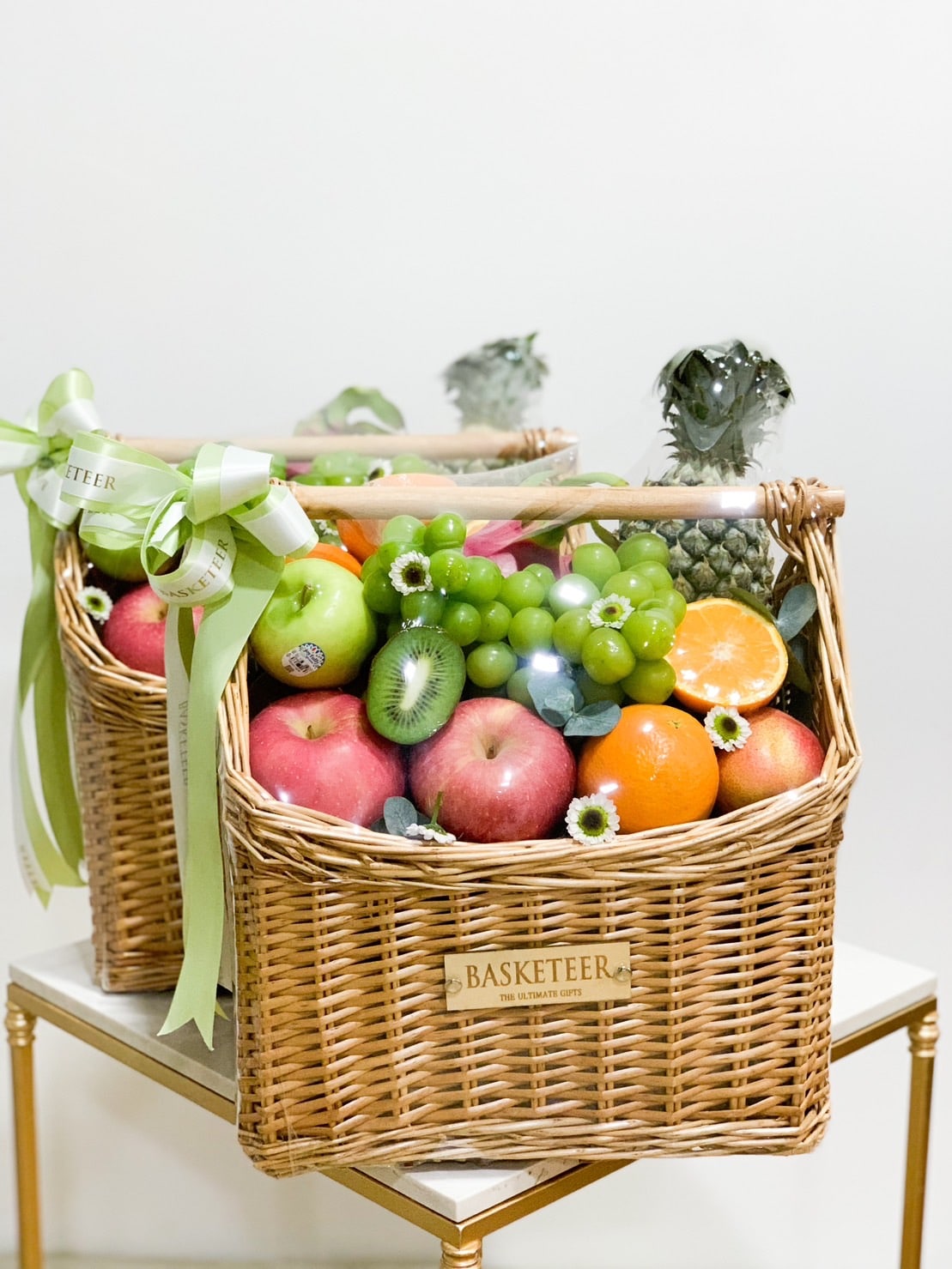 Two wicker baskets filled with various fruits, including apples, kiwi, oranges, grapes, and pineapples, are placed side by side on a table. Each delightful fruit hamper has a green ribbon tied to the handle and a label that reads 