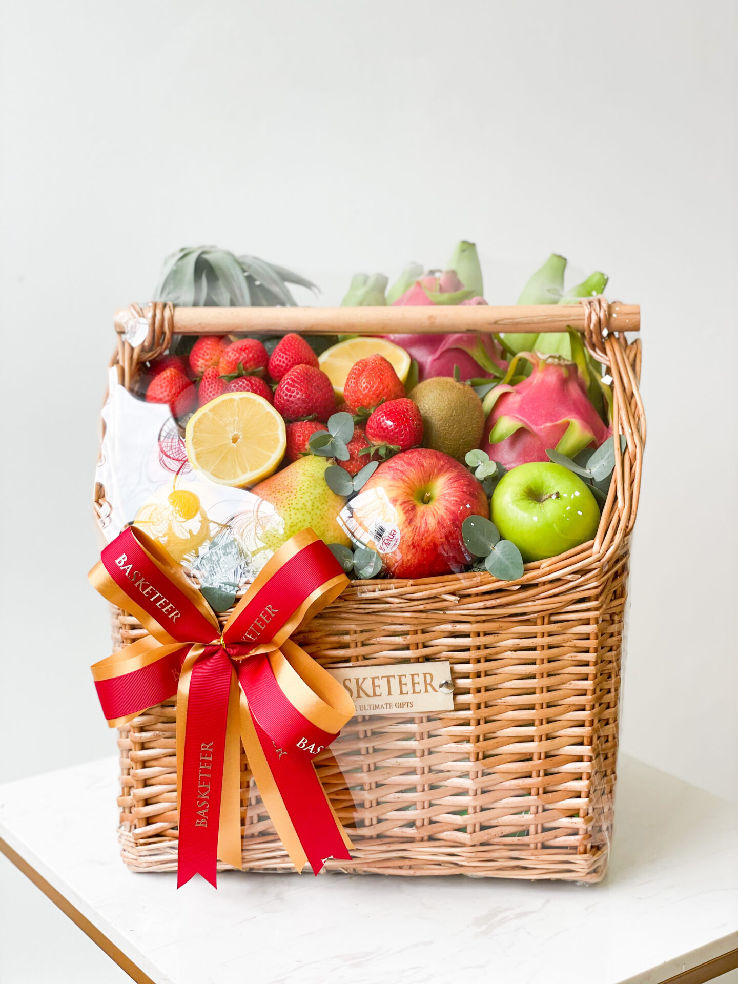 An opulent fruit gift basket filled with assorted fresh fruits including strawberries, apples, dragon fruits, and a halved lemon. The woven basket features a red and yellow ribbon tied in a bow with the brand name 