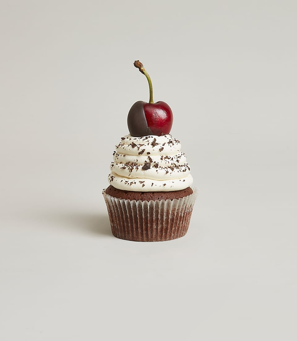 A decadent chocolate cupcake adorned with luxurious swirls of velvety white frosting, elegant chocolate shavings, and a cherry half-dipped in rich chocolate takes center stage. The exquisite treat sits invitingly against a sophisticated grey backdrop, perfectly embodying the delightful indulgence of Basketeer's renowned Mix And Match Berry Cupcakes. A subtle touch of the Basketeer logo on an elegantly textured ribbon adds a finishing mark of luxury to this gourmet creation.