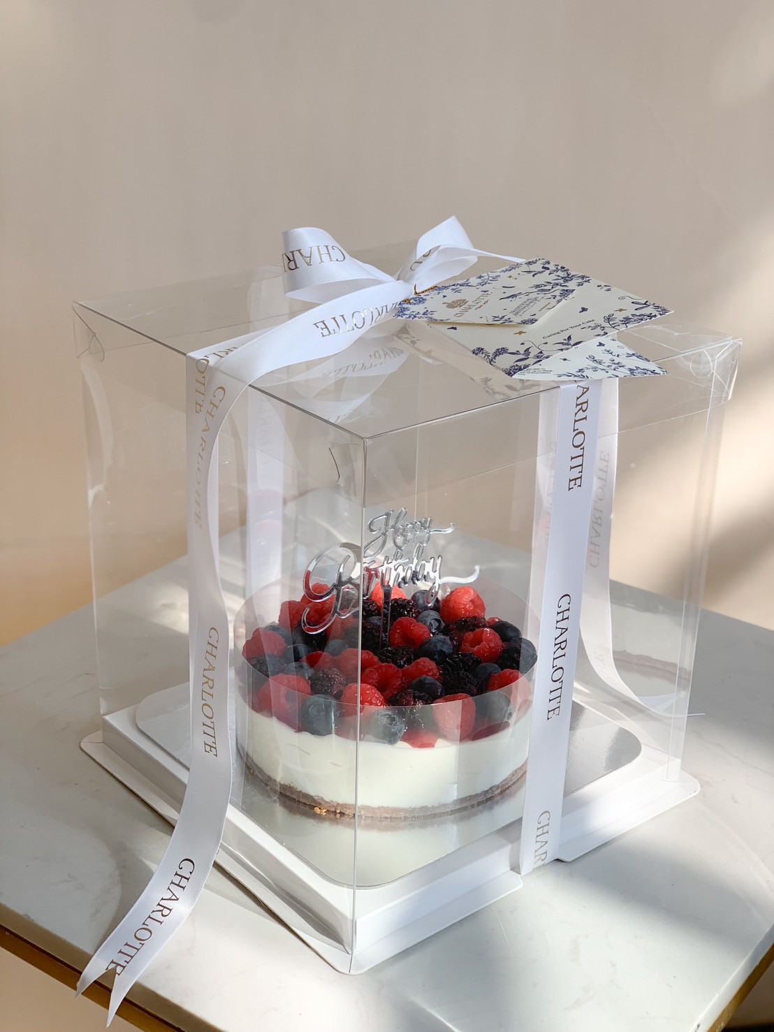 A beautifully packaged mixed berry cheesecake with fresh raspberries, blueberries, and blackberries, presented in a clear gift box with a ribbon.