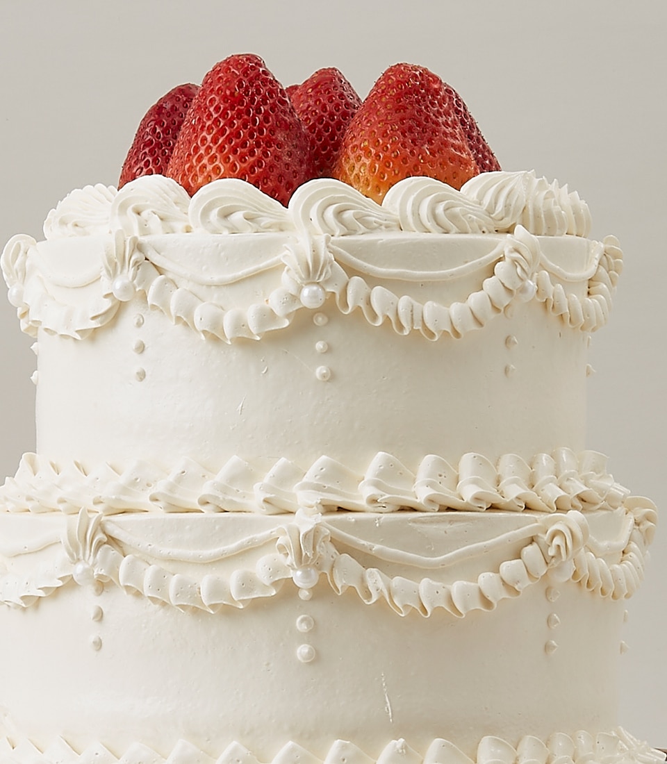 A close-up view of an opulent two-tier white cake, exquisitely adorned with intricate swirls of pristine white icing and delicate pearl-like embellishments. The top tier, fit for royalty, is crowned with luscious whole strawberries that infuse a vibrant pop of red. The neutral backdrop ensures all attention is lavished on this magnificent creation from Basketeer. Nearby, a luxurious box embossed with the Basketeer logo subtly suggests the pedigree and craftsmanship that defines this masterpiece.