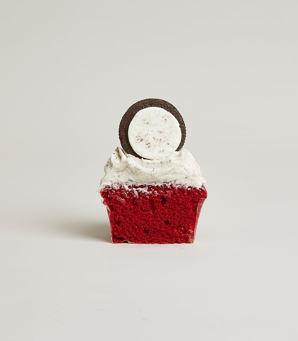 A luxurious red velvet cupcake adorned with a generous swirl of creamy white frosting, crowned with a rich chocolate sandwich cookie meticulously poised vertically. The scene exudes elegance against a pristine white backdrop. The product is presented with utmost sophistication, featuring the Basketeer logo discreetly embossed on a ribbon that adds an extra touch of class to this exquisite dessert masterpiece.