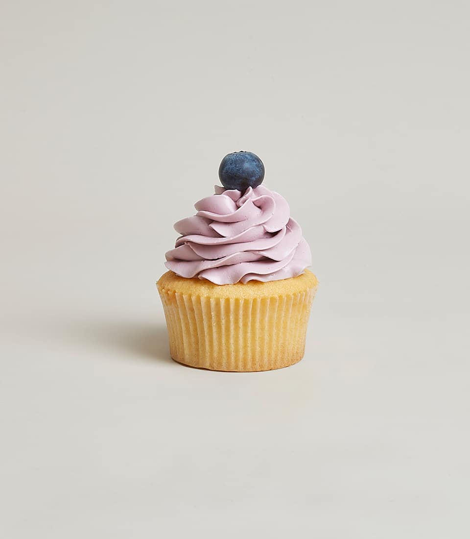 A delectable vanilla cupcake graced with an elegant swirl of light purple frosting and crowned with a single, luscious blueberry rests invitingly on a light gray background. This luxurious confection perfectly captures the essence of Blueberry Belly Cupcakes by Basketeer, effortlessly combining sophistication and indulgence.