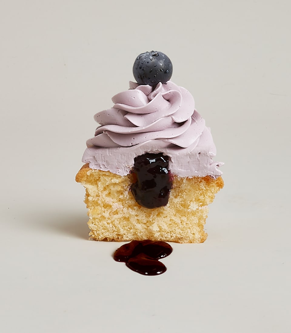 A sumptuous halved Blueberry Belly Cupcake from Basketeer, featuring moist vanilla sponge crowned with rich, velvety purple frosting and adorned with a fresh blueberry. The glossy dark jam filling luxuriously spills onto the pristine white plate, creating a tantalizing display of indulgence. A ribbon and a sophisticated box bearing the Basketeer logo sit elegantly in the background, enhancing the scene with an air of opulence.