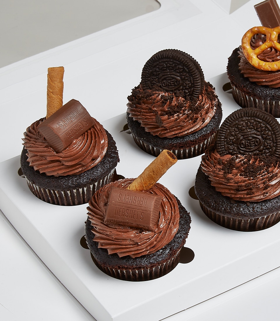 A lavish assortment of Mix & Match Chocolate Cupcakes is elegantly presented in a pristine white box. Each decadent cupcake showcases luxurious swirls of chocolate frosting, adorned with either a sumptuous square of rich chocolate, an indulgent Oreo cookie, or a perfectly salted pretzel. The box bears the distinguished Basketeer logo, adding an extra touch of opulence to this inviting display.