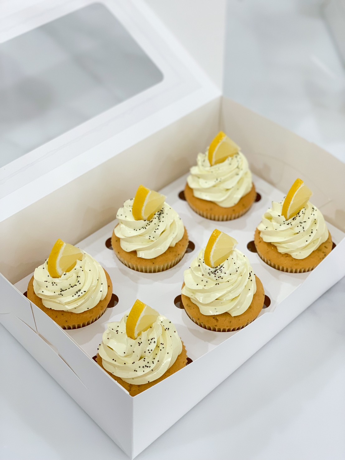 A pristine white box adorned with the Basketeer logo reveals a tempting assortment of six Perfect Poppyseed and Lemon Curd Cupcakes. Each delicacy is crowned with an opulent swirl of velvety white frosting, a tantalizing slice of zesty lemon, and an elegant dusting of poppy seeds. The open box, resting gracefully on a luxurious white marble surface, invites you to indulge in the exquisite flavors crafted by Basketeer.