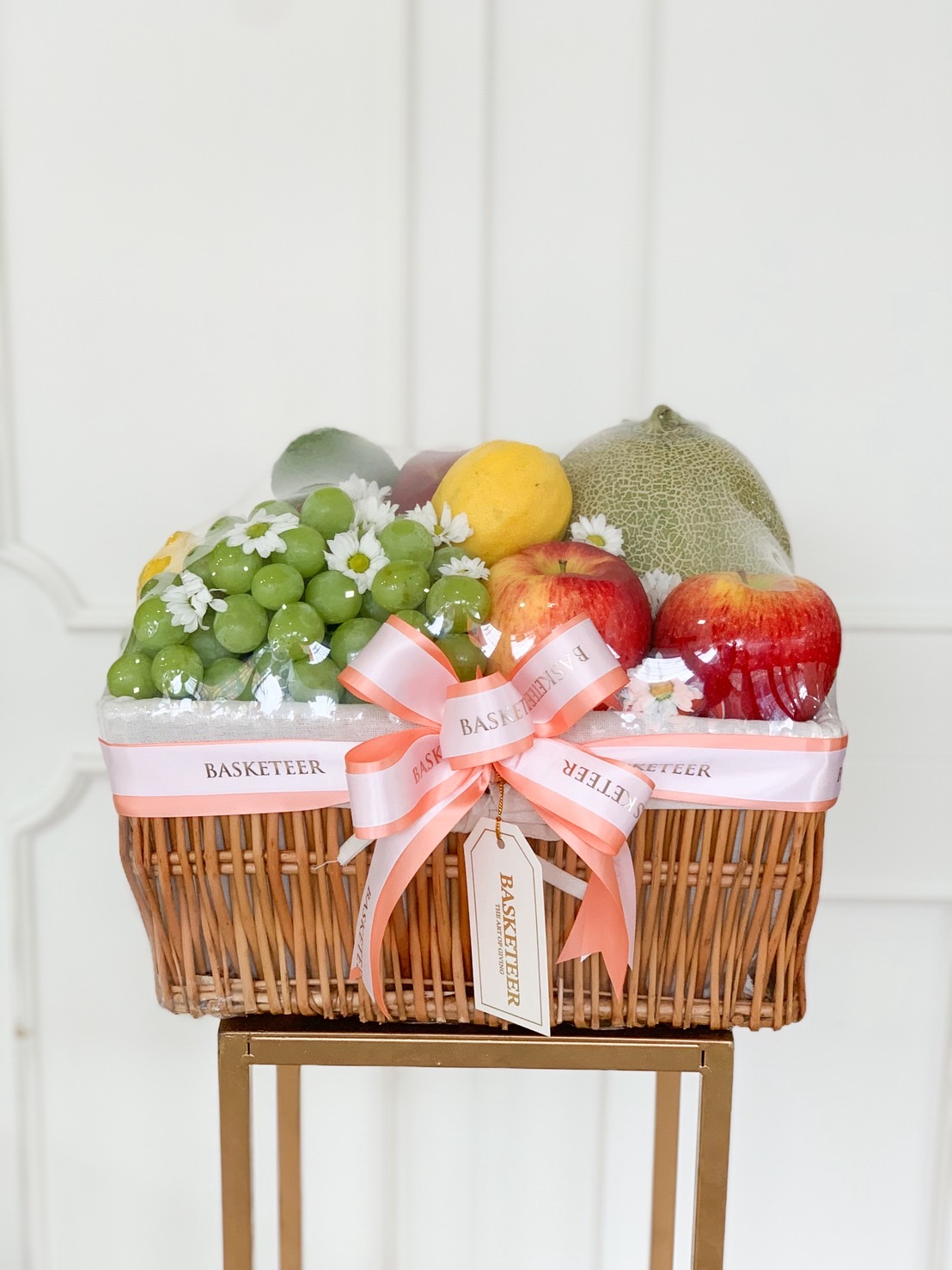 Luxury fruit basket filled with green grapes, red apples, melon, mango, and seasonal fruits, decorated with a peach bow and white daisy flowers.