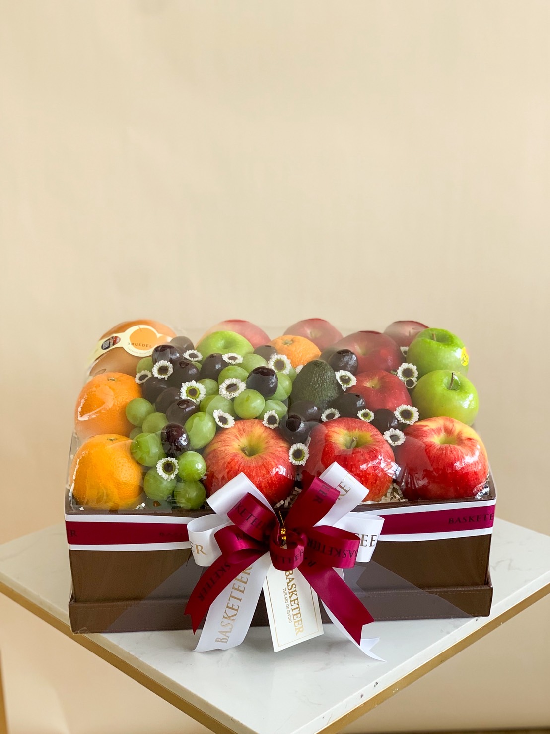 Premium fruit hamper with apples, grapes, oranges, avocados, and fresh flowers in a luxury square box with a burgundy ribbon.