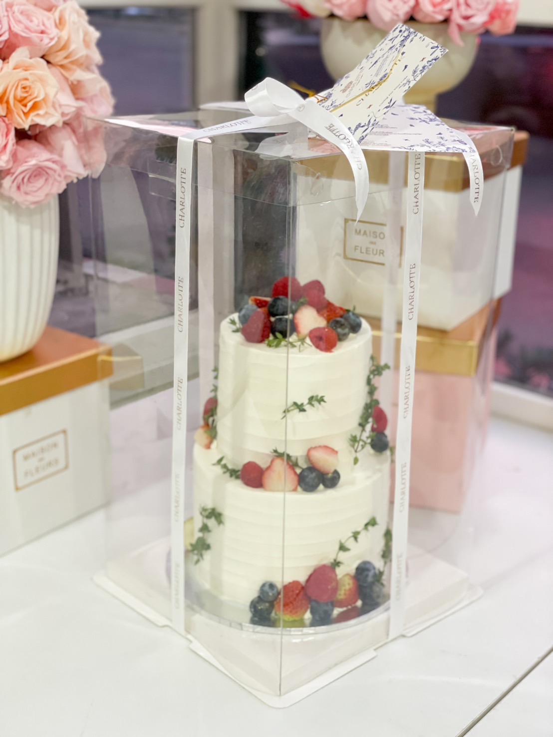 A 2-tier white buttercream cake decorated with fresh berries and greenery, presented in a luxury gift box, perfect for weddings and celebrations.