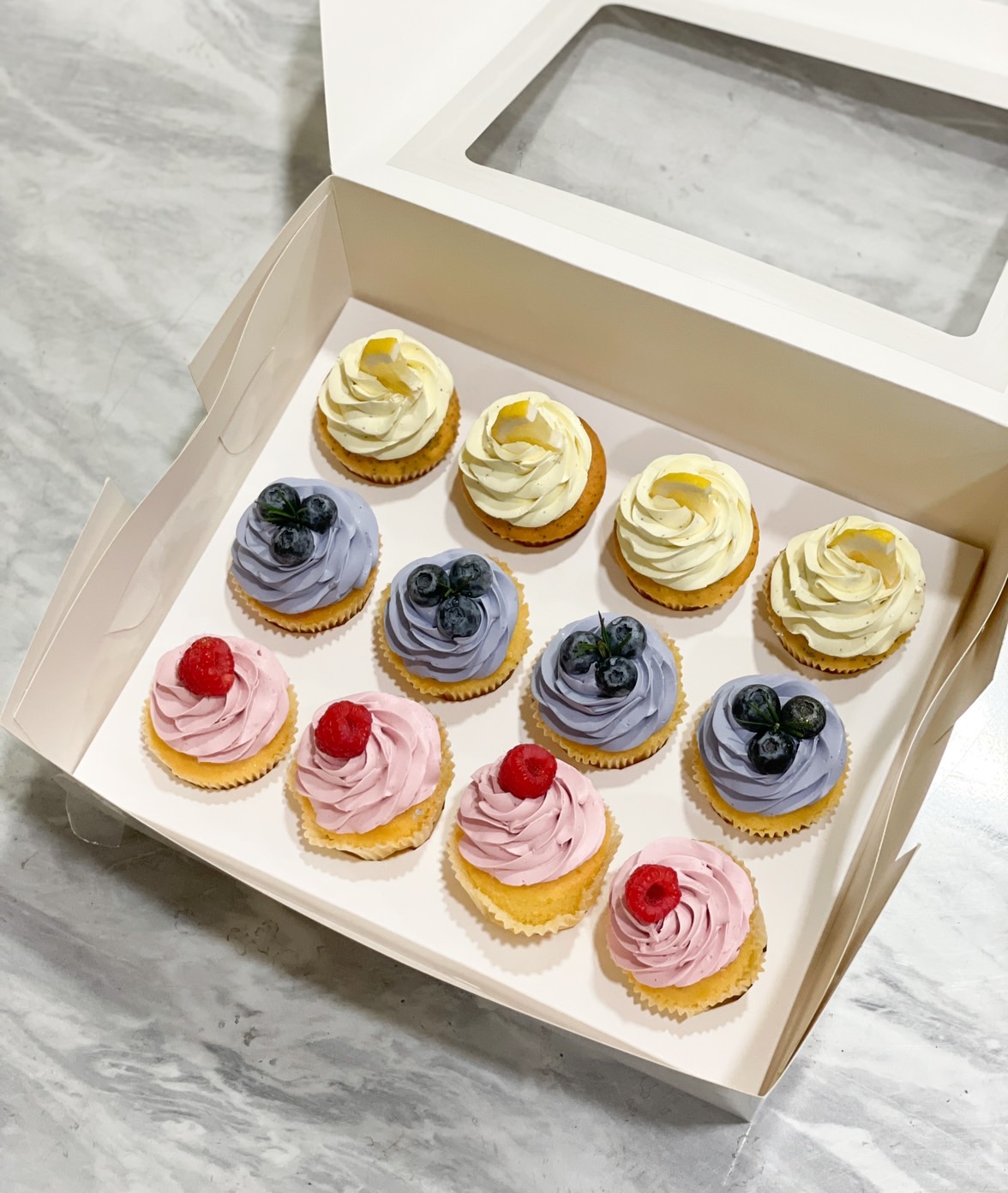 A beautifully arranged box of 12 gourmet cupcakes with blueberry, raspberry, and lemon buttercream frosting, garnished with fresh berries.