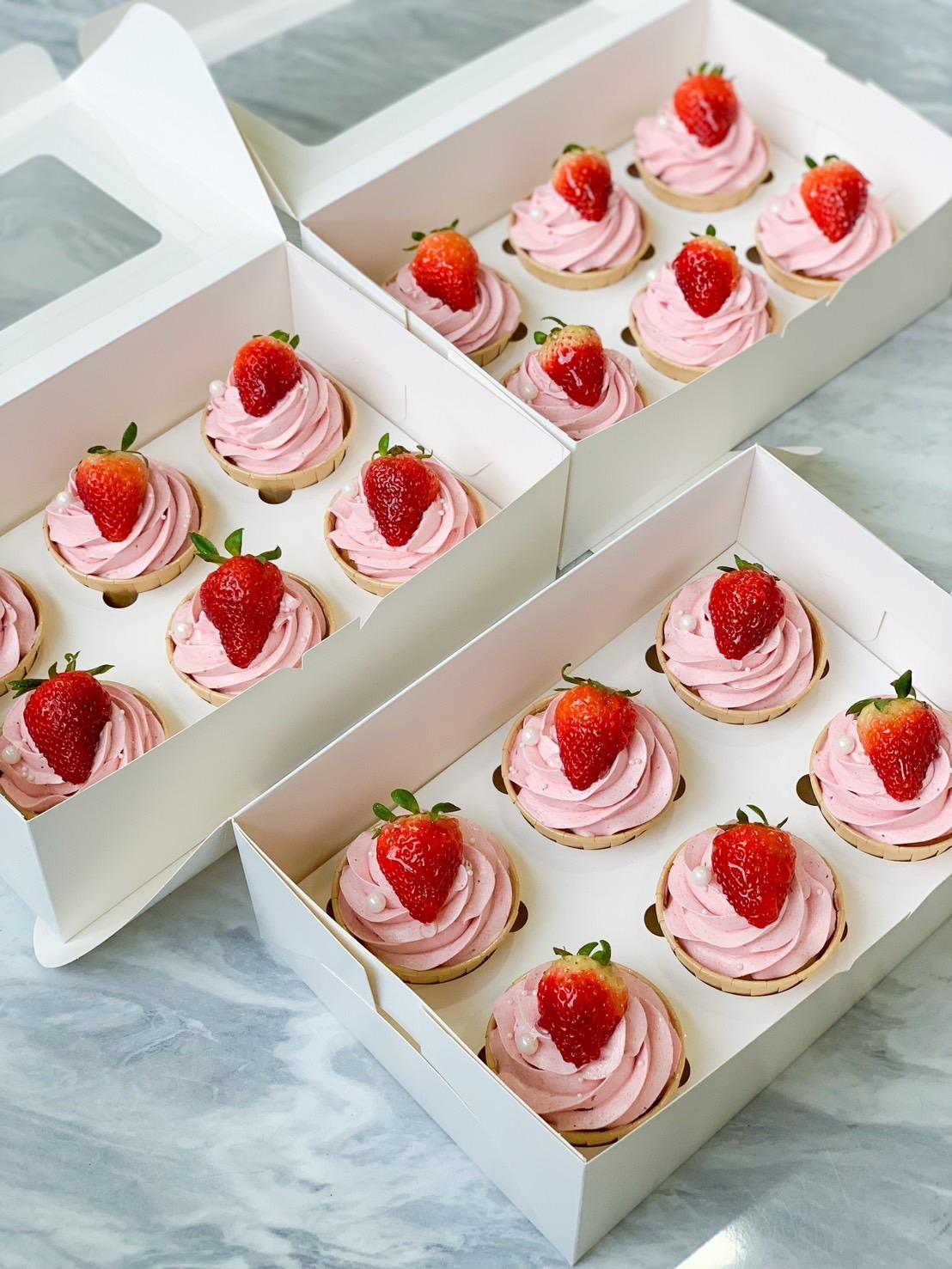Three white boxes filled with Rather Raspberry Jam Centred Cupcakes. Each cupcake is topped with pink frosting, a fresh strawberry, and decorative white pearls. The boxes are arranged on a light gray marble surface, the open lids revealing the beautifully decorated cupcakes inside.