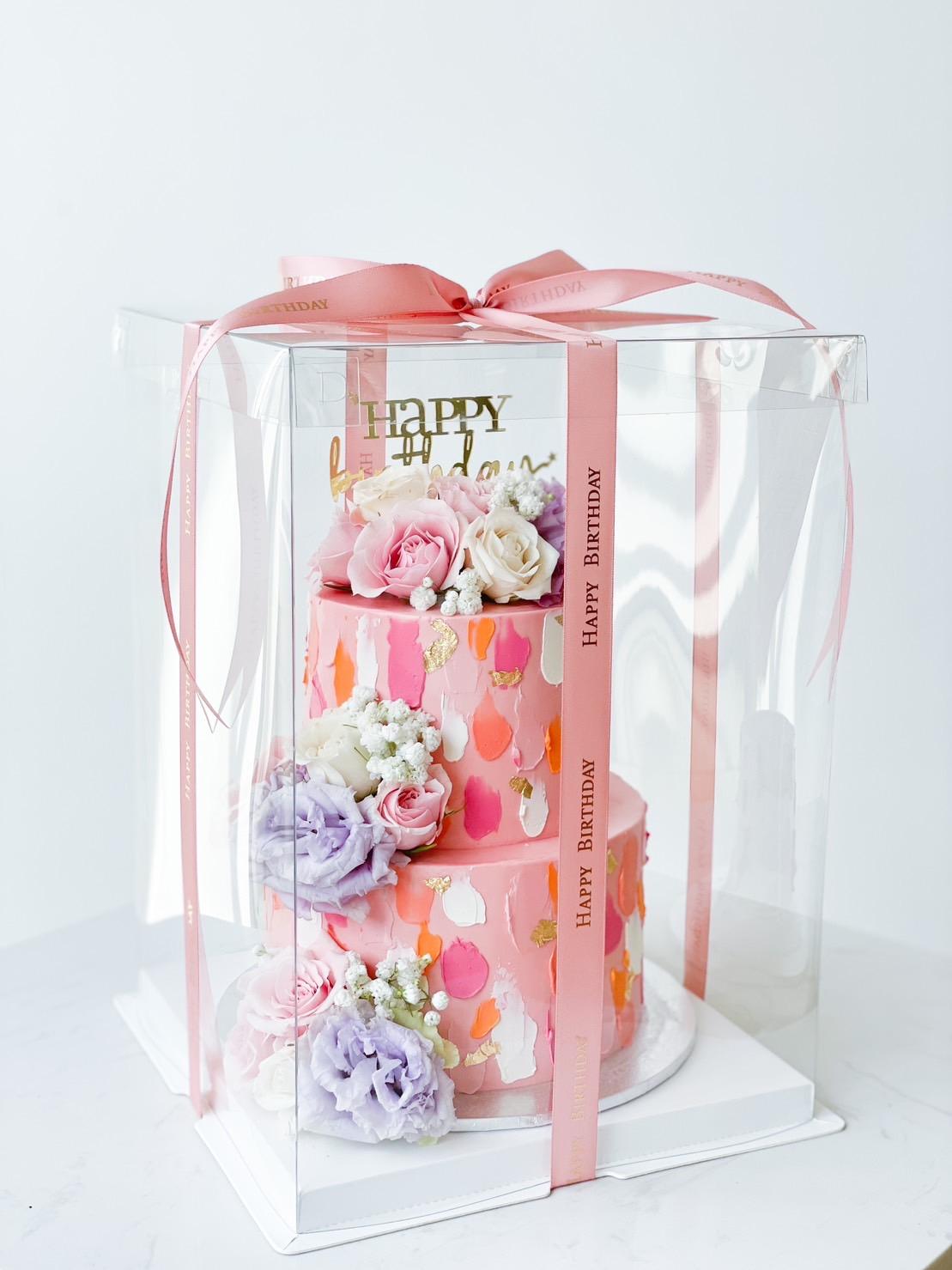 A Bu Cake with 2-Tier Blooms, elegantly decorated with white, pink, and purple flowers, is enclosed in a clear plastic box. 