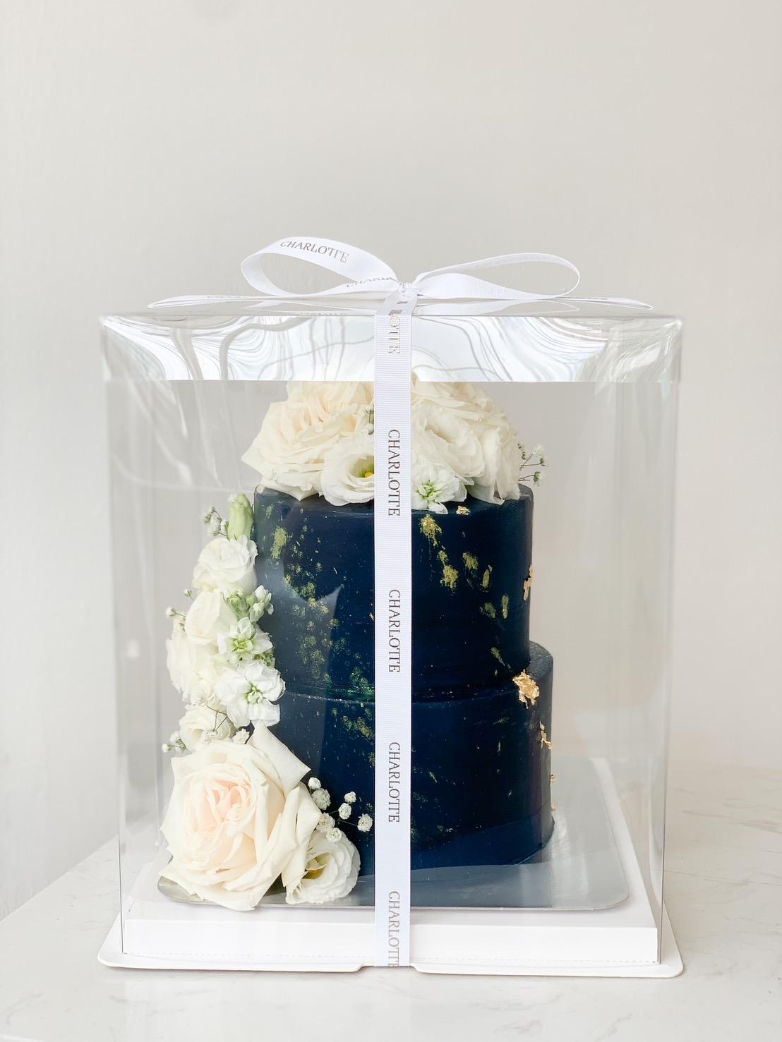 A sophisticated two-tier dark blue Charlotte cake adorned with white roses, presented in an elegant clear box with a white ribbon.
