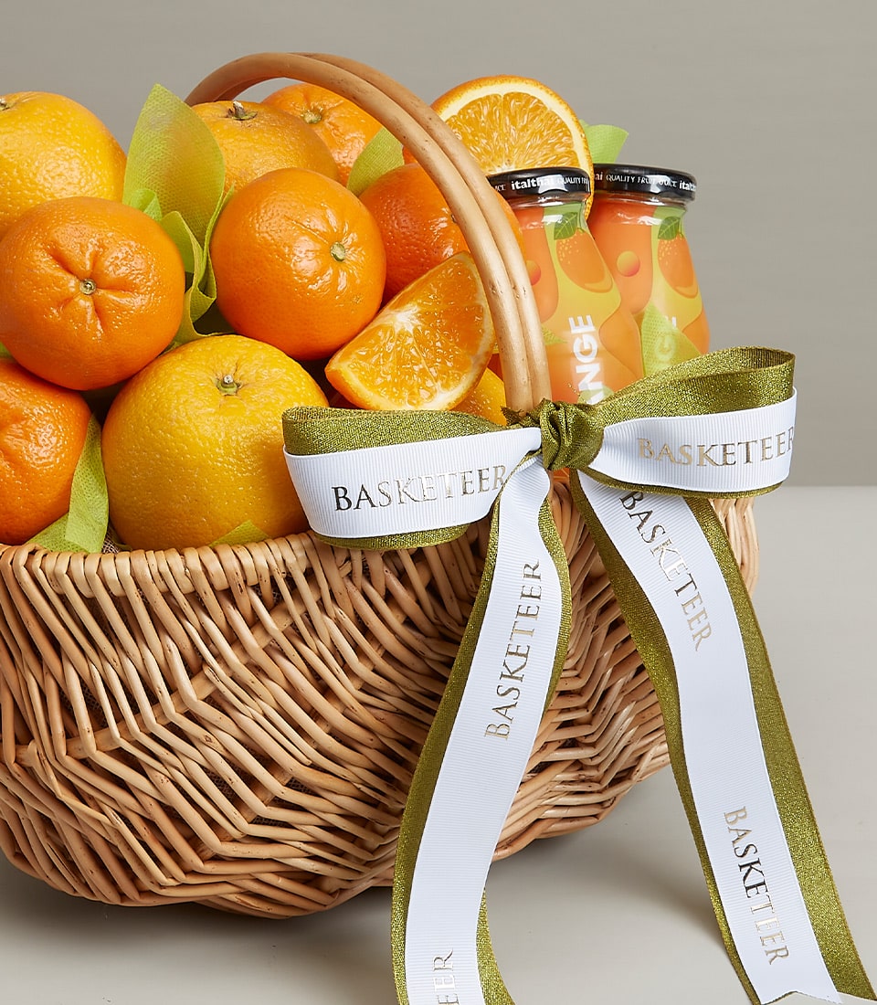 A wicker basket filled with fresh oranges and orange slices, accompanied by two bottles of orange juice. The basket has a green and white ribbon with the word 