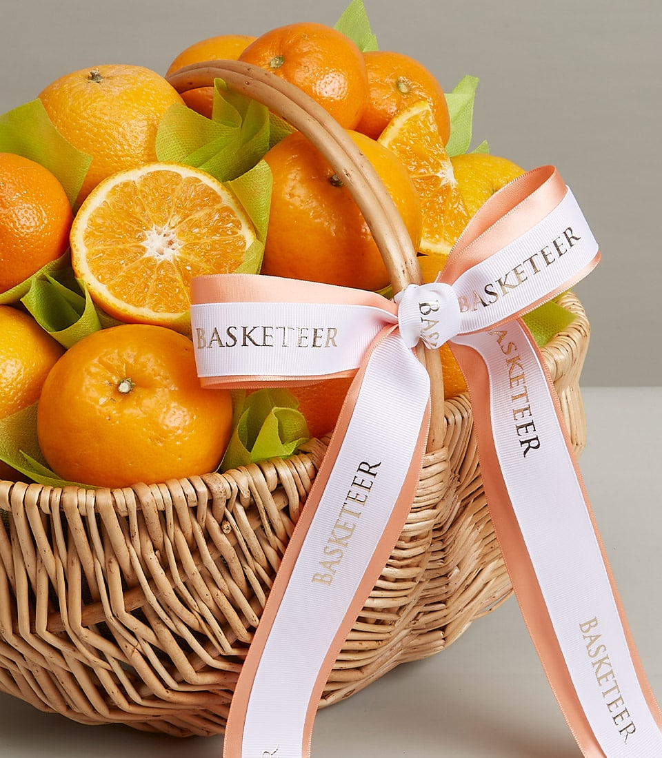 A luxurious wicker basket from Basketeer, brimming with fresh, vibrant oranges, including some whole and one exquisitely sliced in half. Elegantly adorned with a sophisticated white and peach ribbon proudly showcasing the 