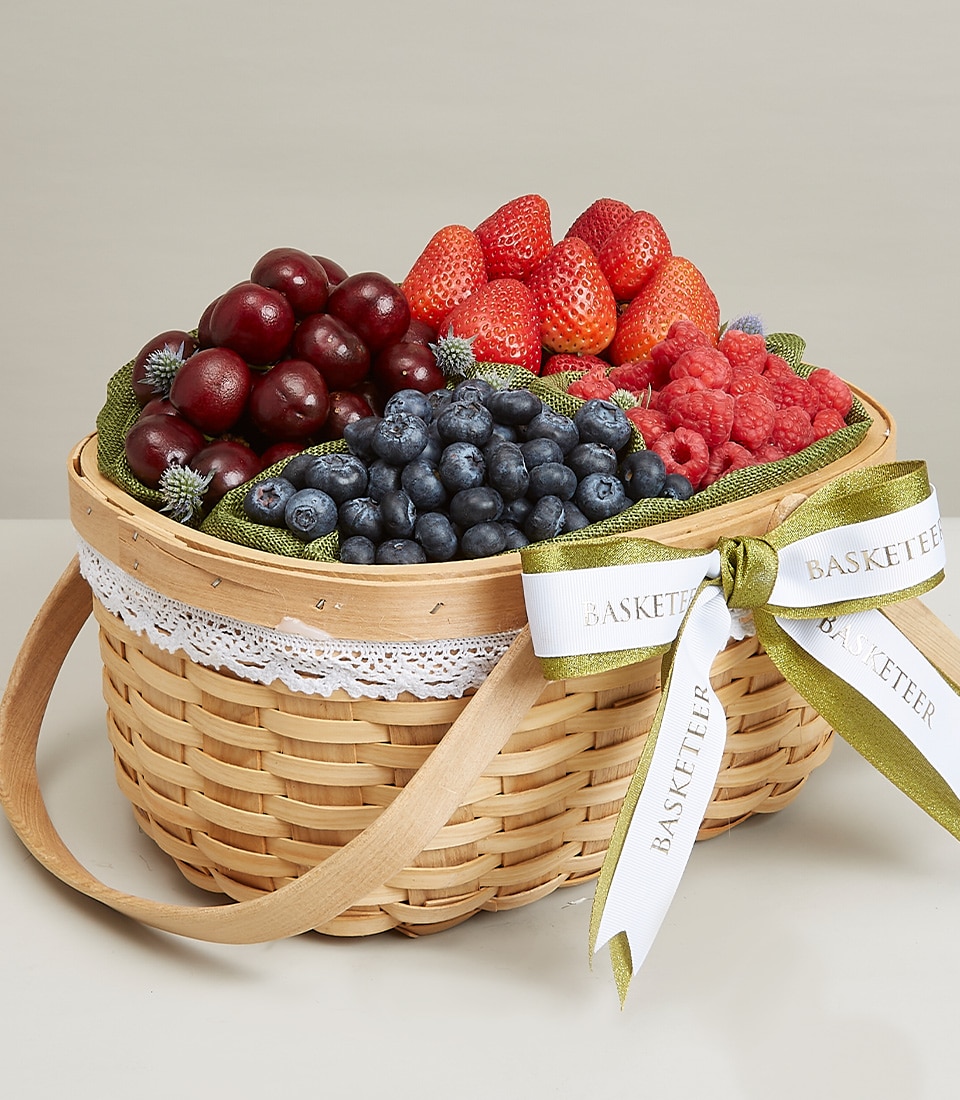 A luxurious woven basket overflowing with an exquisite selection of fresh cherries, strawberries, blueberries, and raspberries, each jewel-like berry adorned with delicate green leaves. This opulent Berry Orchard treasure chest is elegantly finished with a decorative white lace trim and an enchanting green and white ribbon proudly bearing the 