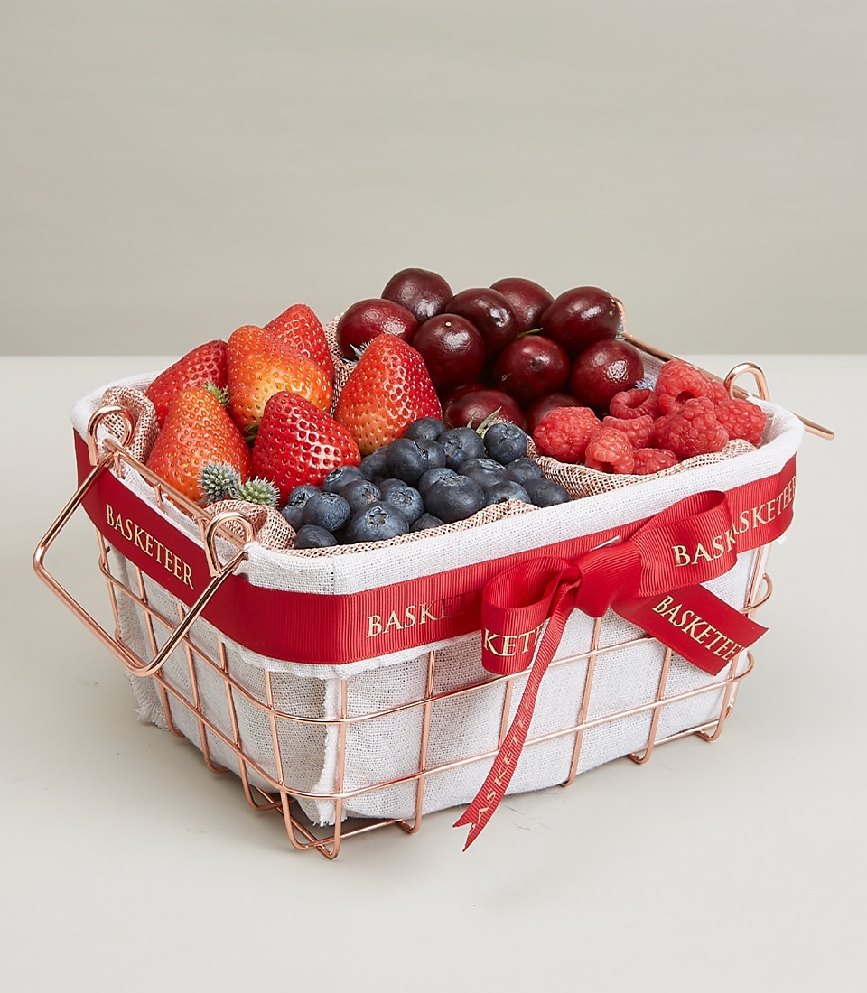 A opulent mixed berry basket from Basketeer, this exquisite copper wire creation is elegantly lined with pristine white cloth, showcasing an array of luscious fresh fruits—succulent strawberries, plump grapes, vibrant blueberries, and juicy raspberries. The basket is tastefully adorned with a luxurious red ribbon bearing the prestigious 