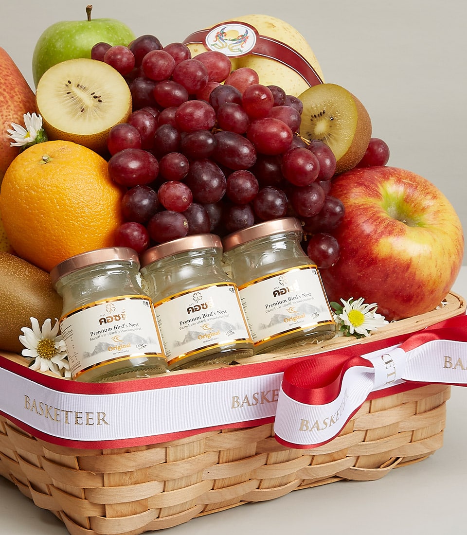 An elegantly woven basket brimming with an exquisite selection of fresh grapes, apples, oranges, and kiwis. Tucked thoughtfully among the luscious fruits are three premium jars of honey, promising a blend of natural sweetness and luxury. Enhancing its sophisticated allure is a pristine white ribbon with vibrant red edges, adorned with the illustrious 