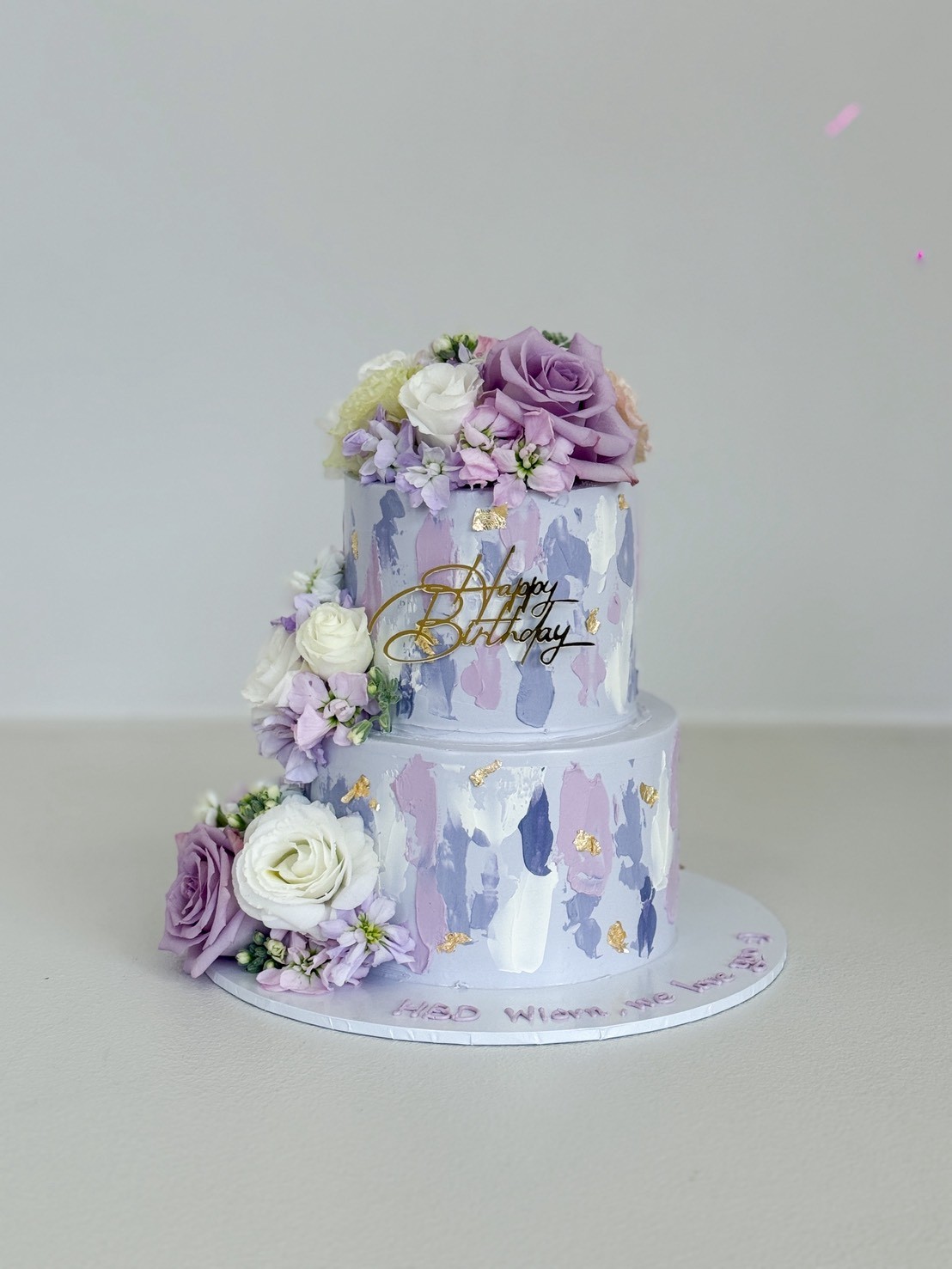 Lavender two-tier floral cake decorated with fresh roses and lilac brush strokes – luxury birthday cake design in pastel tones.