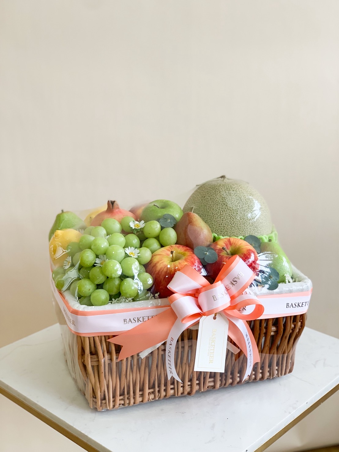 Premium fruit basket with Japanese melon, green grapes, apples, pears, and assorted fresh fruits in a wicker basket with peach ribbon.