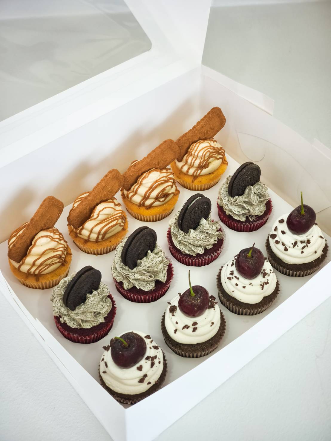 A Charlotte Bakery white box reveals nine fairy cakes, elegantly topped with caramel, Oreo biscuits, or whipped cream and cherries.