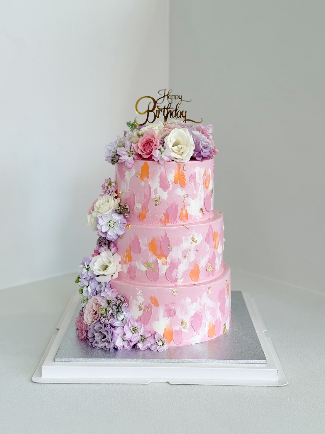 Three-tier pink birthday cake with pastel brushstroke design and fresh flowers in pink, purple, and white hues.