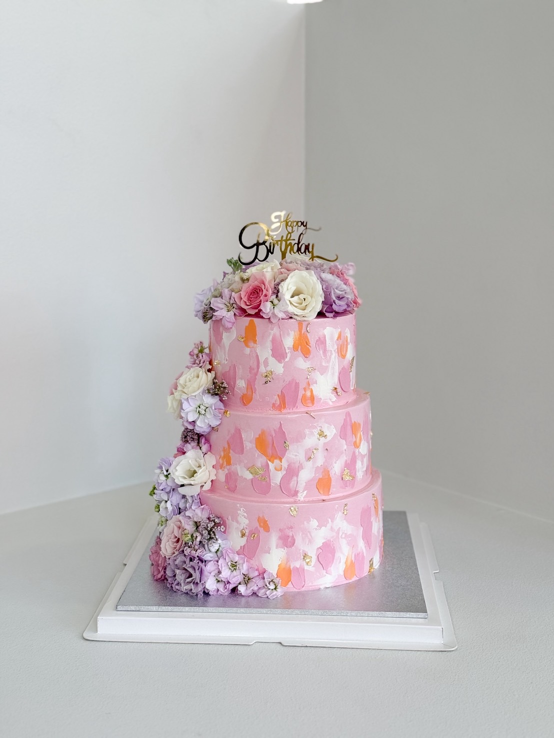 Three-tier pink birthday cake with pastel brushstroke design and fresh flowers in pink, purple, and white hues.