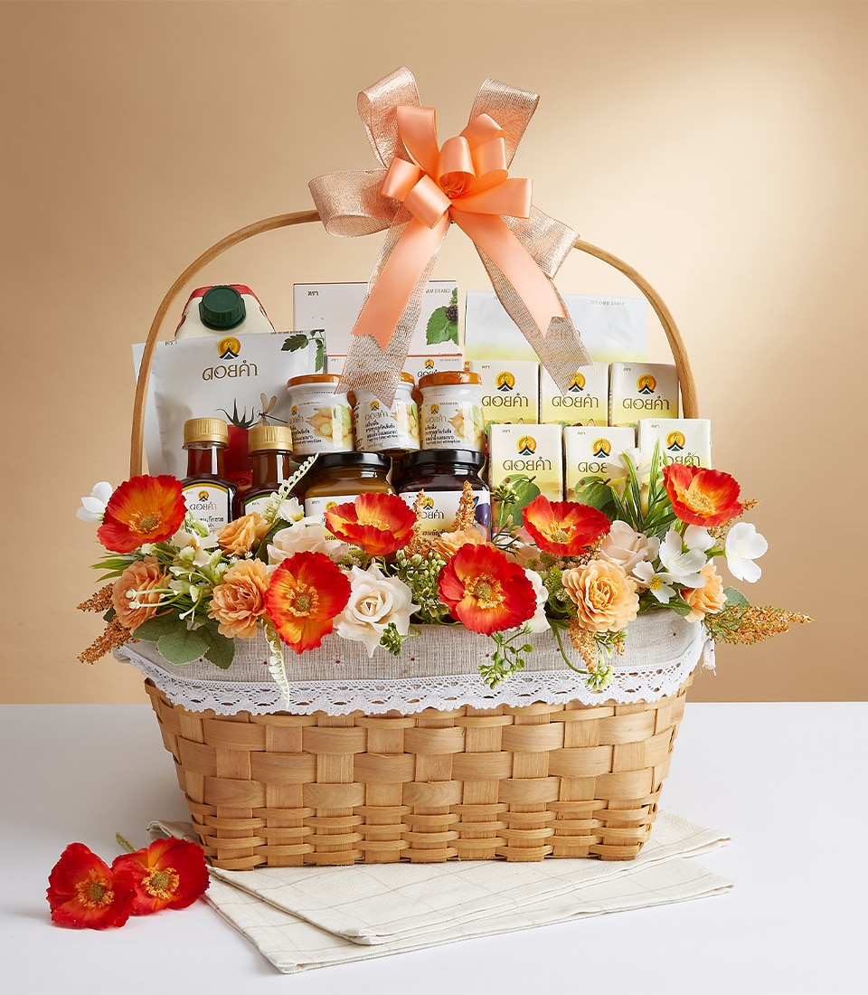 A luxurious wicker basket, lined with an elegant lace-trimmed white cloth, brimming with an assortment of gourmet packaged delicacies and artisanal jars. The arrangement is exquisitely embellished with vibrant orange and red flowers, and crowned with a lavish orange ribbon bow bearing the Basketeer logo. All set against a warm beige backdrop, this scene exudes sophistication and invites indulgence.