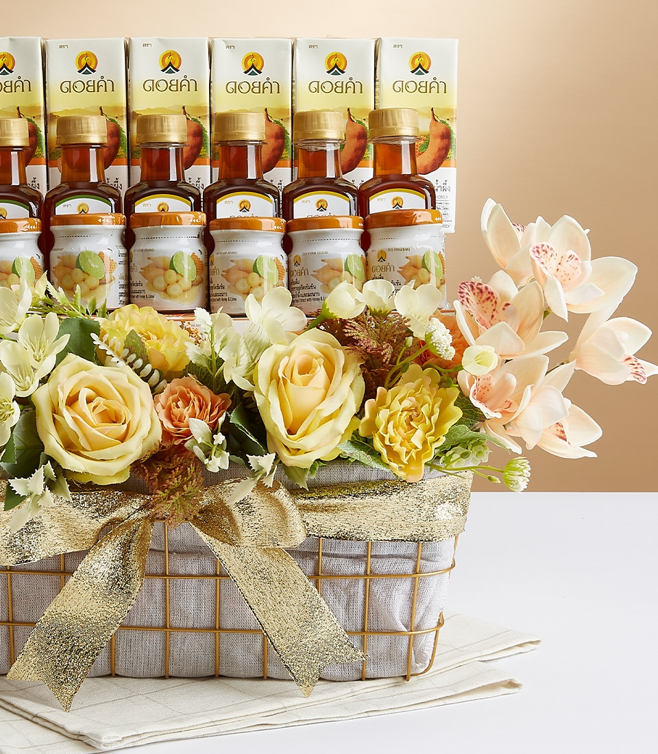 A lavish Basketeer decorative basket, elegantly adorned with gold ribbons, overflows with vibrant yellow roses and lush greenery. Perched above the radiant blooms are several exquisite small bottles, each boasting a label with a charming lemon graphic. A striking pink orchid branch artistically extends from the right side of this opulent arrangement, creating a scene of unparalleled sophistication and beauty.