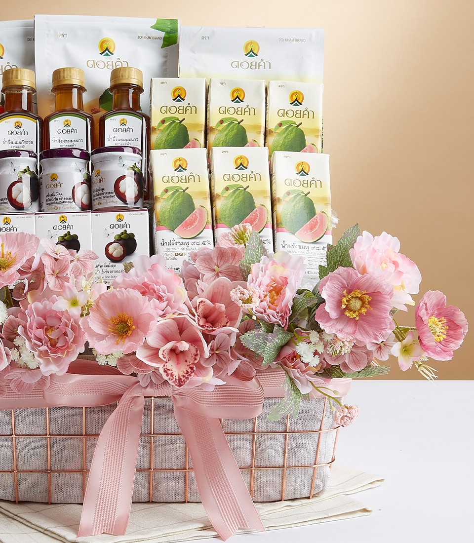A luxurious decorative basket elegantly adorned with delicate pink flowers and silk ribbons showcases an assortment of opulent treasures. Nestled within are several bottles filled with a rich, dark liquid, each one proudly bearing a golden logo and intricate Thai script. Accompanying them are exquisite packets of drink mixes or teas, also embellished with the same golden emblem. The scene is completed by the presence of the Basketeer logo gracing a sumptuous ribbon or box, adding an extra touch of sophistication to this inviting display.