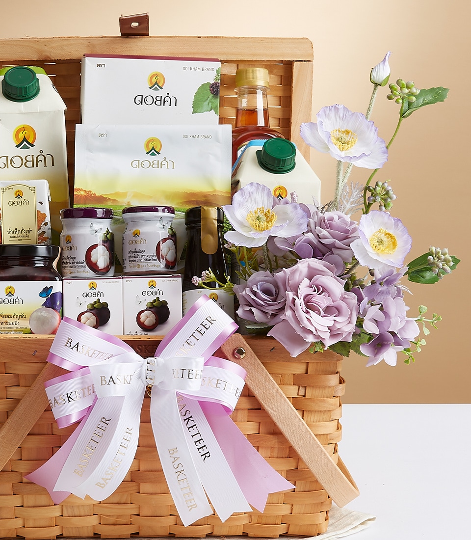 A luxurious wicker gift basket brimming with an exquisite selection of gourmet snacks and beverages, elegantly presented in jars and cartons. The basket is tastefully adorned with delicate lavender and white flowers, and tied with enchanting white and pink ribbons gracefully showcasing the 