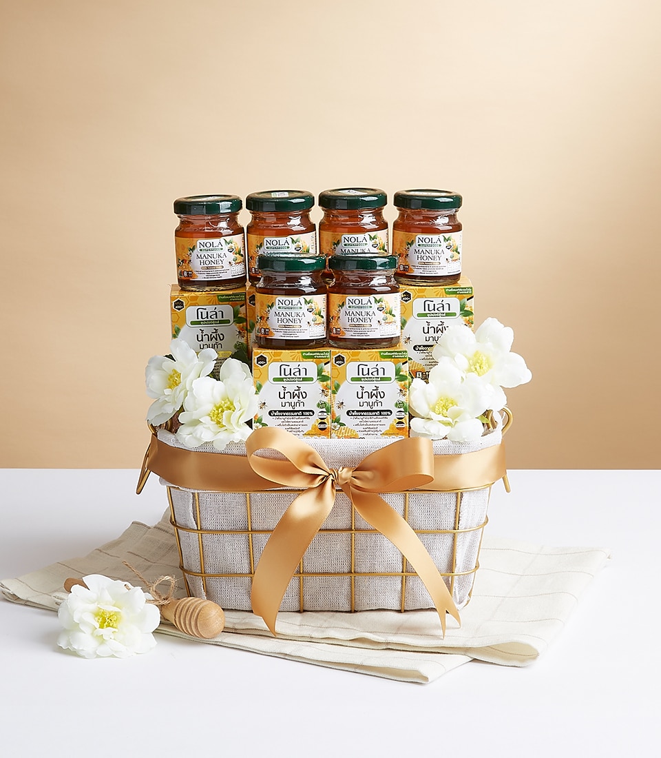 A luxurious wicker basket graced with elegant white flowers and a sophisticated golden bow features an exquisite collection of Nola Honey jars. Nestled upon a pristine white cloth within the basket, the beautifully arranged jars underscore a sense of refinement and indulgence. In front of this opulent display sits a dainty honey dipper and an additional delicate white flower, all poised on a light table, perfectly capturing Basketeer's commitment to elegance and quality in every detail.
