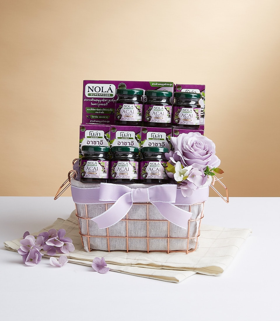 A luxurious gift basket elegantly adorned with a light purple Basketeer ribbon, featuring six exquisite jars of NOLA Acai Berry Essence. The presentation is enhanced by a decorative purple rose and scattered petals, all beautifully arranged on a pristine folded white cloth, set against an inviting beige backdrop.
