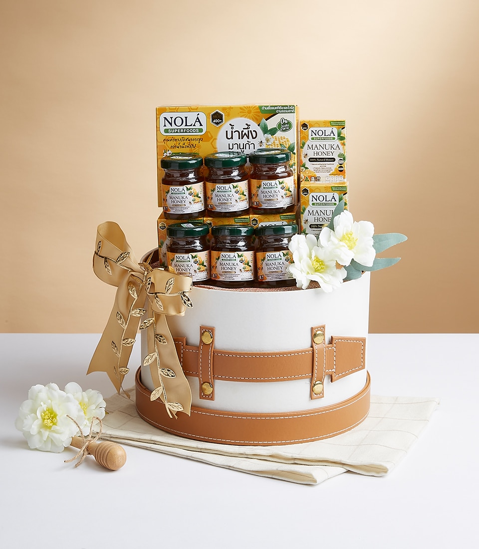 A luxurious white cylindrical gift box featuring elegant tan leather straps is beautifully decorated with a sophisticated beige ribbon and resplendent white flowers. Inside the box, you'll find an exquisite selection of jars filled with premium NOLA Manuka Honey. The lavish packaging proudly displays the Basketeer logo, signaling exceptional quality and care. This opulent gift set is tastefully arranged on a pristine white cloth, accompanied by a charming wooden honey dipper and an additional delicate white flower—creating a captivating scene that exudes luxury and invites indulgence.