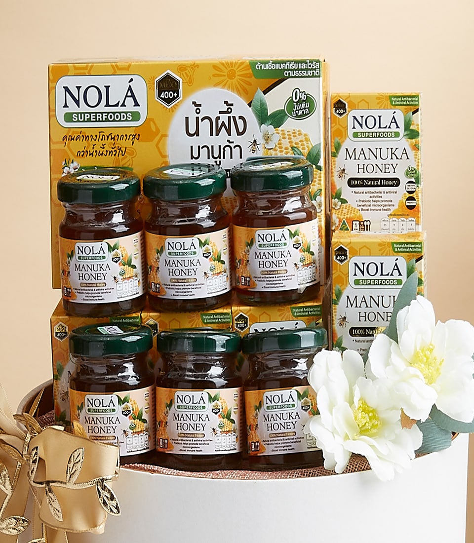 A sumptuous display of Nola Superfoods Manuka Honey products exudes elegance and indulgence. Luxurious jars of the finest honey are artfully arranged in front of a sophisticated Basketeer box, each adorning exquisite honeycomb and floral designs. The Basketeer logo subtly enhances the richness of the presentation. To complete this enchanting scene, a pristine white bloom with lush green leaves gracefully accompanies the opulent honey delights, inviting you to savor nature's most exquisite luxury.
