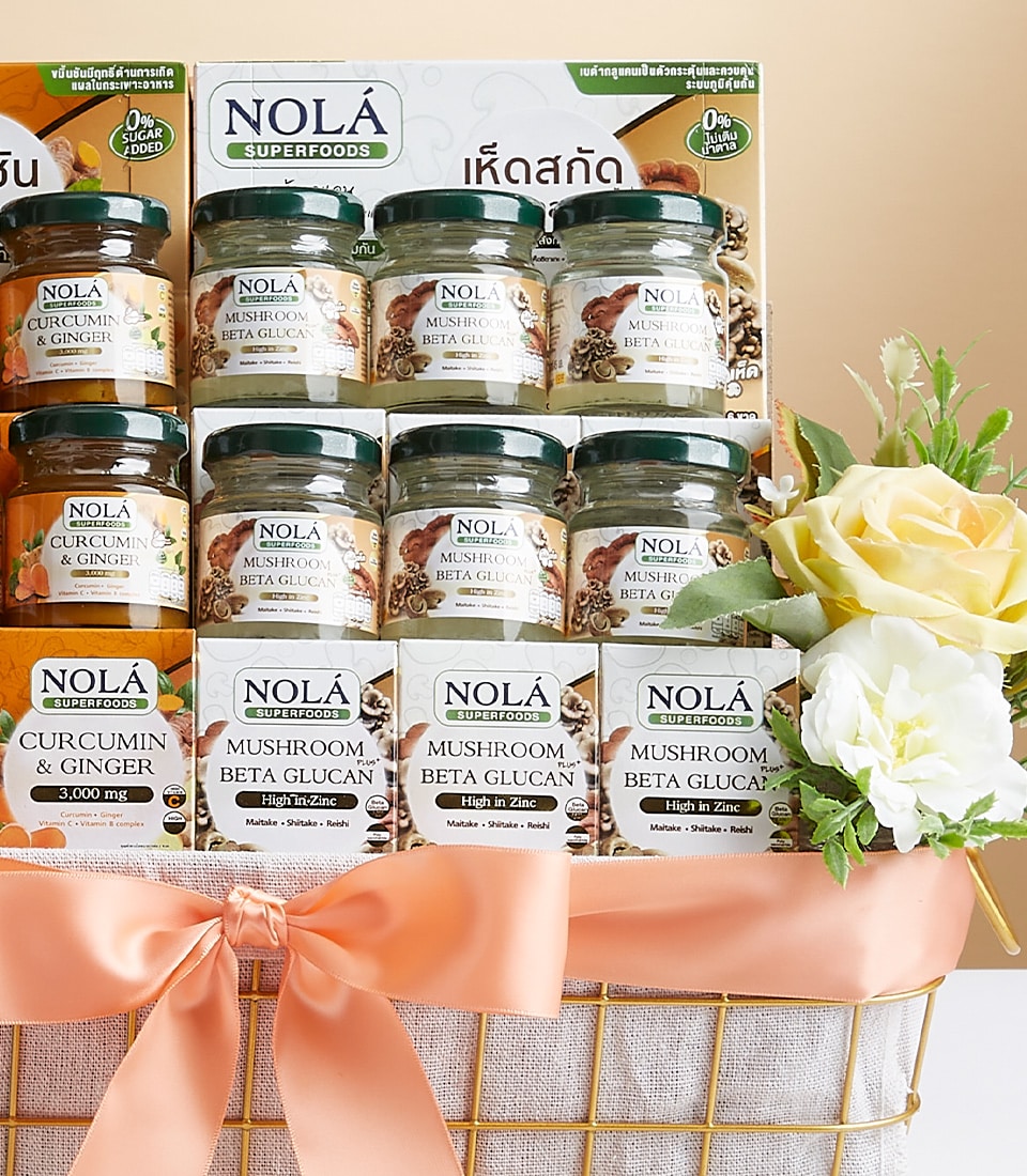 An opulent gift basket, elegantly adorned with a peach-colored ribbon bearing the distinguished Basketeer logo, showcases an exquisite selection of NOLA Superfoods jars. Inside, you'll find premium products such as Curcumin & Ginger and Mushroom Beta Glucan+. The basket is further embellished with a stunning bouquet of yellow and white roses on the right side, creating a scene of luxury and wellness.