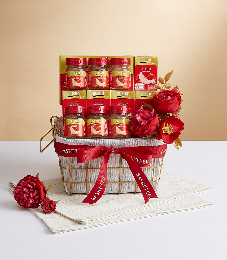 An exquisite gift basket from Basketeer, beautifully adorned with luxurious red ribbons bearing the Basketeer logo. Nestled within are premium bottles of Nestlé Musashi bird's nest drink, meticulously arranged to captivate and delight. The basket is elegantly decorated with lush red and peach-colored flowers that exude opulence. The serene background features a soft gradient of beige tones, enhancing the refined aesthetic, while the entire arrangement rests gracefully on a pristine white cloth.