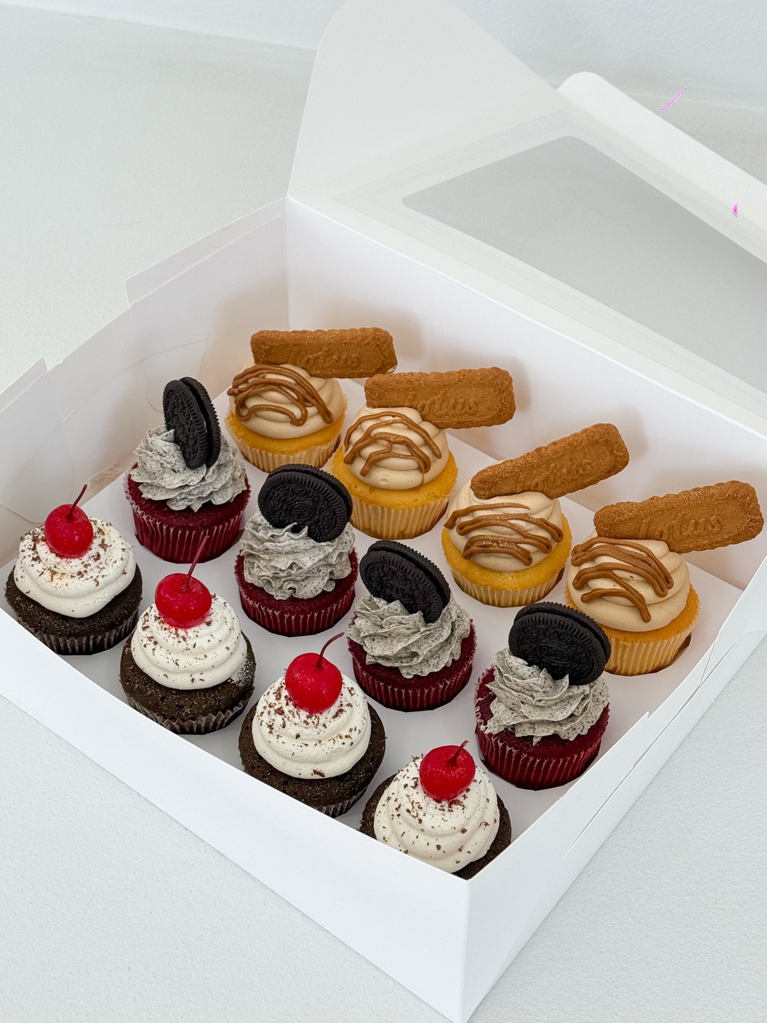 A white box filled with Mix and Match Cupcakes: four chocolate cupcakes topped with cherries, four red velvet cupcakes with Oreo halves, and four cheesecake cupcakes crowned with caramel drizzle and a rectangular biscuit.