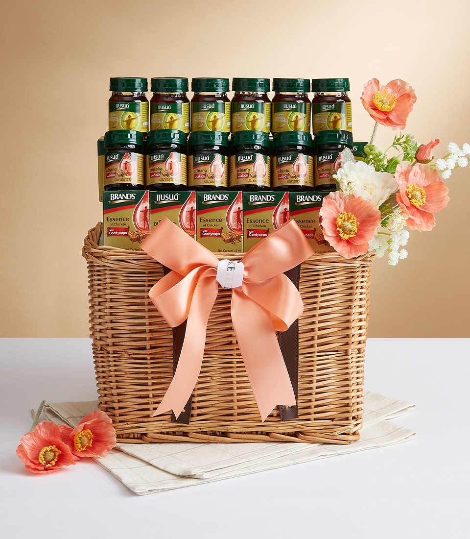A luxurious wicker gift basket elegantly showcases six bottles of Brand's Essence of Chicken, each promising wholesome nourishment. The basket is adorned with a sophisticated orange ribbon and a delicate white tag, hinting at the thoughtful curation within. Draped in exquisite pink and white flowers, this charming arrangement rests atop a soft beige cloth on a pristine white surface, set against an inviting beige backdrop. The scene radiates warmth and refinement, heightened by the presence of the Basketeer logo subtly integrated into the presentation.