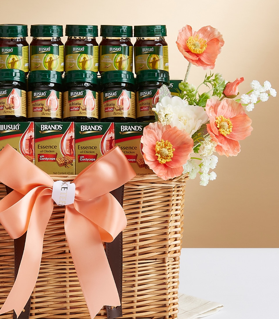 A lavish wicker basket brimming with an array of premium juice bottles, elegantly adorned with a generous peach-colored bow, proudly bearing the Basketeer logo, rests gracefully on a pristine white surface. Accompanying this opulent gift is a stunning bouquet of vibrant orange poppies and delicate white flowers, exquisitely arranged against a soothing beige backdrop. The scene exudes sophistication and warmth, capturing the essence of luxury gifting by Basketeer.