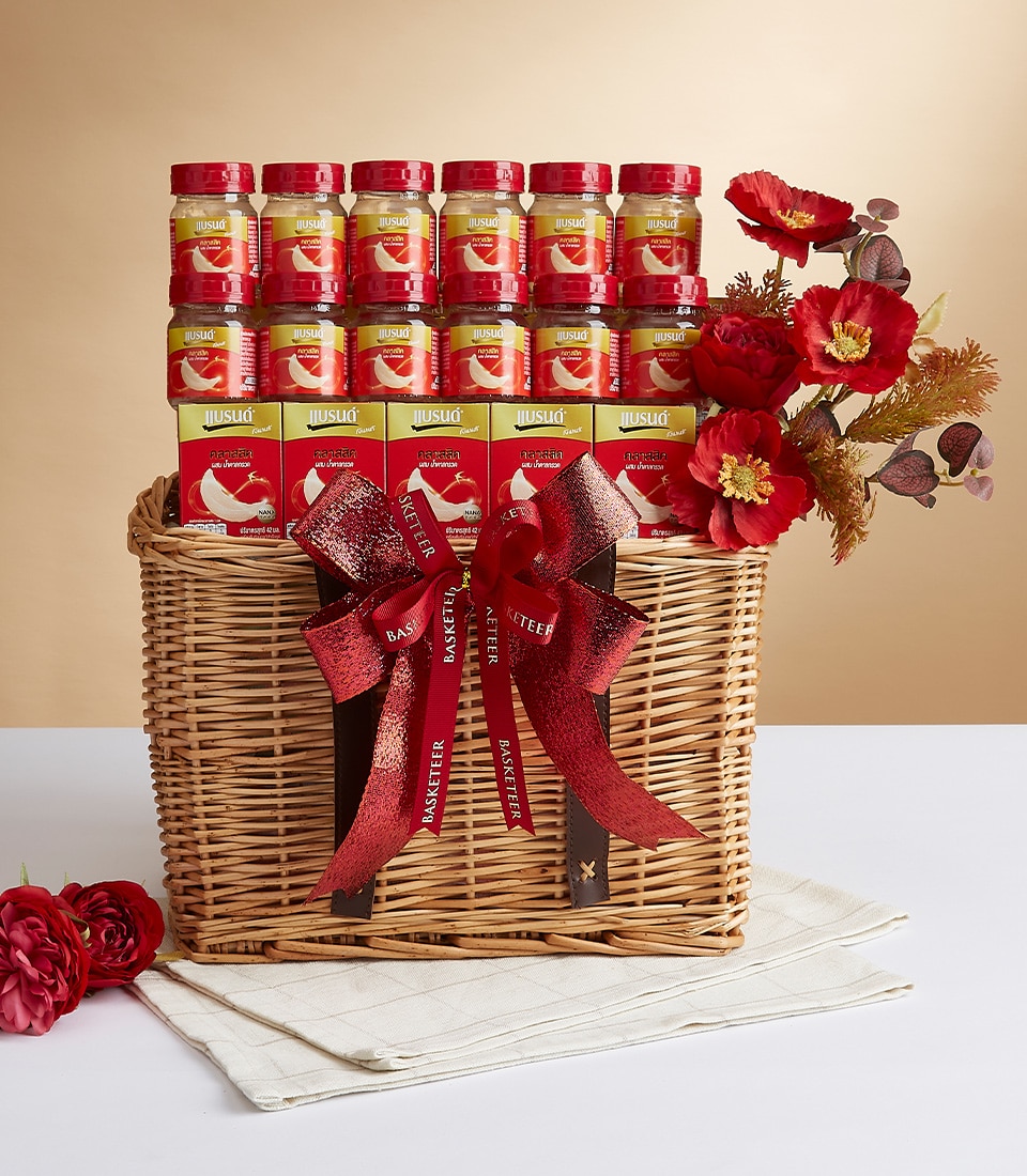 A luxurious wicker basket brimming with an exquisite selection of Basketeer’s premium jars, adorned in elegant red and gold packaging. Lavishly tied with opulent red and gold ribbons bearing the Basketeer logo, this stunning ensemble rests gracefully on a pristine white cloth accented by a delicate red flower. The backdrop features a soft gradient beige, enhancing the sophistication of the scene.
