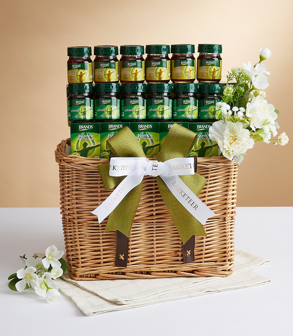 A luxurious wicker gift basket elegantly adorned with a sophisticated green and white ribbon, proudly showcasing the Basketeer logo. The basket is filled with an exquisite selection of Brand's premium chicken essence jars, perfectly arranged to capture attention. Delicate white flowers adorn the basket, enhancing its charm and sophistication as it rests gracefully on a pristine white surface against a warm beige background. The scene is further enriched with scattered white blossoms around the base, creating an inviting and opulent ambiance.