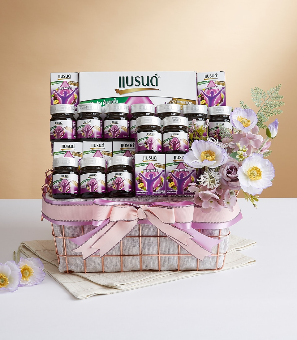An exquisitely curated Basketeer gift basket featuring neatly arranged glass jars with elegant purple labels, beautifully accentuated by a bouquet of pink and white flowers. The basket is adorned with a luxurious lavender ribbon tied in an opulent bow, proudly showcasing the Basketeer logo. This sumptuous ensemble rests on a pristine folded white cloth, creating a serene and sophisticated contrast against the beige backdrop.