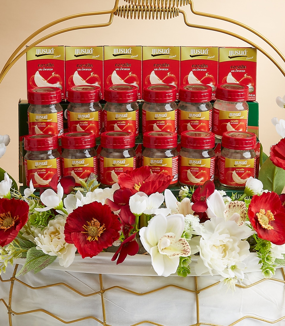 An opulent display of Juslig Bird's Nest beverages graces a tiered, golden stand, each small glass jar adorned with elegant red labels. The luxurious arrangement is further enhanced by a beautiful blend of red and white flowers at the base, creating a sophisticated and inviting presentation. A ribbon tied to the stand proudly showcases the Basketeer logo, adding an exclusive touch to this exquisite ensemble.